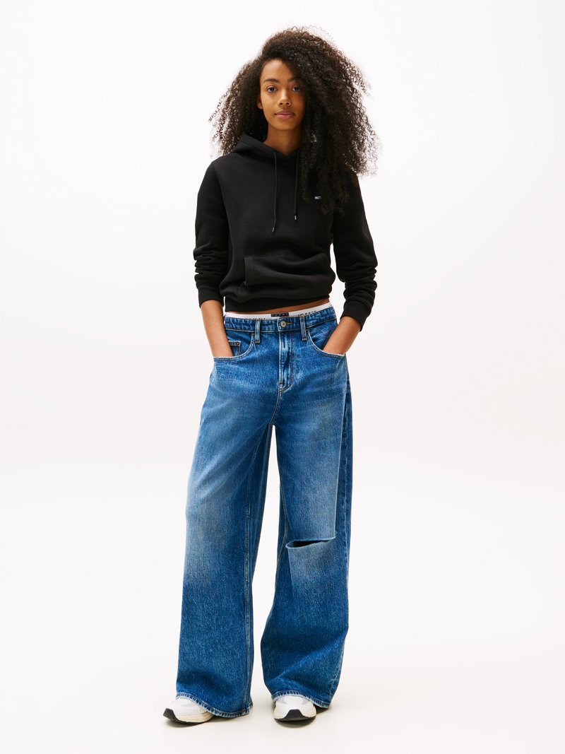 Svart hettegenser, cropped passform; blå vide jeans, falmet, med utskjæringsdetalj; brukt med hvite og svarte sneakers.