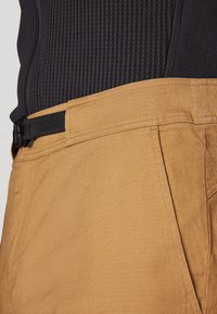 Tan pants made of textured fabric with a smooth finish, featuring a black adjustable belt, side pockets, and subtle stitching details.