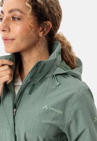 Giacca da pioggia verde chiaro, con texture, dotata di zip frontale e cappuccio regolabile. Presenta un logo sul petto e un motivo di tessuto distintivo.