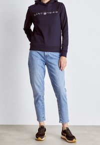 Granatowy hoodie z białym napisem "GANT 1949", w połączeniu z jasnoniebieskimi dżinsami oraz czarnymi sneakersami z żółtymi akcentami i teksturowanymi podeszwami.