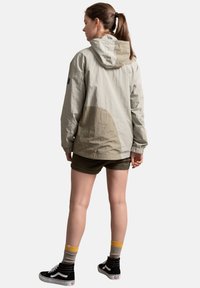 Giacca a vento grigia chiara con accenti beige, caratterizzata da una vestibilità comoda, maniche lunghe e tessuto testurizzato. Indossata con pantaloni corti scuri e sneakers nere.