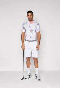 Blomstermønstret polo skjorte i hvit, med blå og rosa detaljer, sammen med hvite shorts og grå belte. Holder en golfkølle.