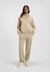 Felpa beige con cappuccio e tasca a marsupio, abbinata a pantaloni tapered coordinati e sneakers beige chiaro con dettagli testurizzati.