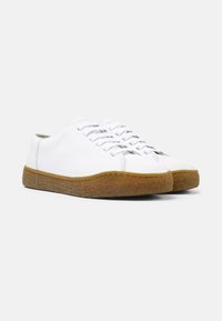 Sneakers bianchi in pelle con suola in gomma marrone chiaro e texture, caratterizzati da un design semplice e sei coppie di fori per i lacci.