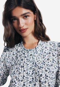 Weiße Bluse mit floralen blau- und grünmuster, gerafftem Ausschnitt und gefältelter Vorderseite. Weicher Stoff mit langen Ärmeln und floralen Akzenten.