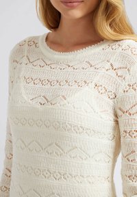 Pull léger de couleur crème avec un motif tricoté comprenant des designs en dentelle horizontaux et diagonaux. Encolure ronde et manches longues.