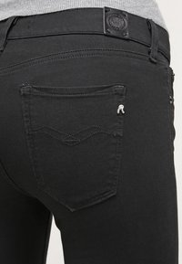 Jeans de mezclilla negra con un diseño ajustado, que cuentan con un patrón de costura distintivo en el bolsillo trasero y una etiqueta con el logo en la cintura.