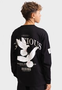 Zwart langemouwen shirt met witte duiven en tekstgraphics op de achterkant, gemaakt van zachte stof met een relaxte pasvorm.