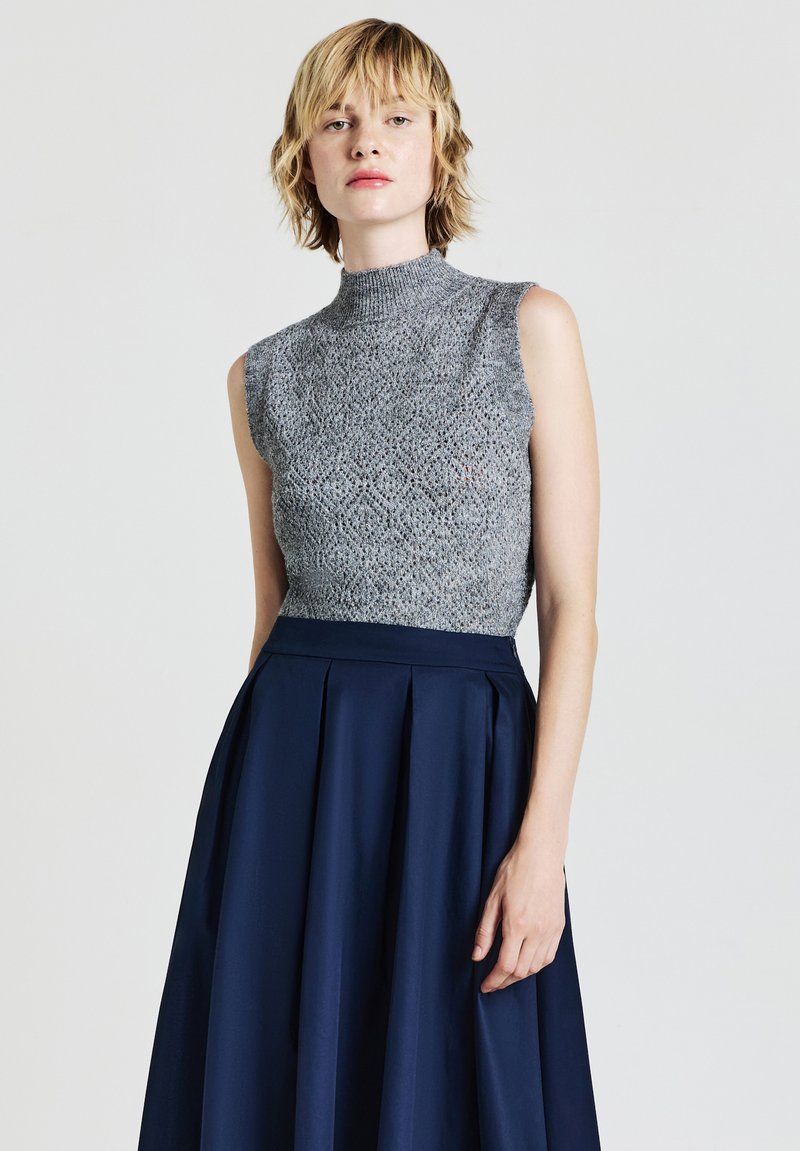 Graues ärmelloses Stricktop mit hohem Kragen, kombiniert mit einem marineblauen Faltenrock. Das Top zeichnet sich durch ein strukturiertes, offen gewebtes Muster aus.