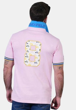 Homme aux cheveux foncés portant un polo rose avec un col bleu, des poignets de manches rayés, et un grand numéro 8 avec des motifs de voitures dans le dos.