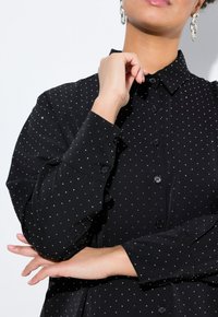 Camicia nera con bottoni a maniche lunghe, caratterizzata da un motivo a pois argentati. Maniche lunghe con polsini a bottone. Tessuto liscio e design del colletto classico.