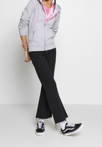 Sudadera gris con cremallera y mangas largas sobre una camiseta rosa con estampado tie-dye, combinada con jeans negros acampanados y zapatillas negras con detalles blancos.