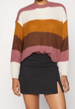 Frau trägt einen langärmeligen, gestreiften Strickpullover in Weiß, Senf, Braun und Mauve, kombiniert mit einem schwarzen Minirock mit einem vorderen Oberschenkelschlitz.