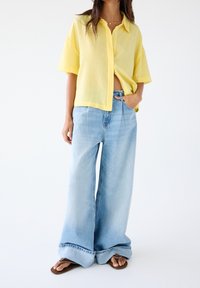 Chemise jaune en lin à manches courtes avec col, associée à un jean en denim bleu clair à jambes larges et des tongs marron. Coupe décontractée et ample.