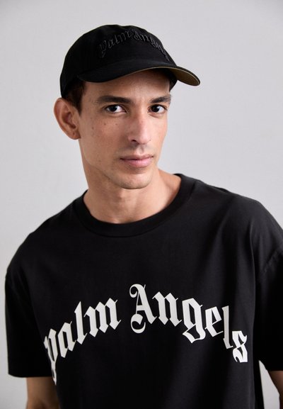 Black t-shirt featuring bold white "Palm Angels" text in a Gothic font, paired with a black cap with matching embroidery, set against a neutral background.