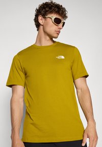 T-shirt od pamučne tkanine u gorućoj žutoj boji s kratkim rukavima, s malim bijelim logotipom na prsima. Glatka tekstura, dizajn s okruglim izrezom.