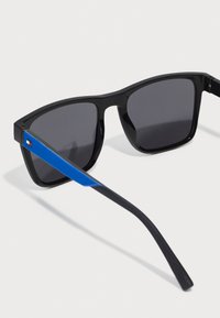 Ochelari de soare negri cu lentile gri, având o dunga albastră pe tâmple. Acest cadru are un design elegant, unghiular.