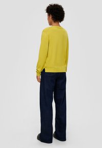 Gelber Strickpullover mit rundem Halsausschnitt und gerippten Bündchen, kombiniert mit dunkelblauen weit geschnittenen Jeans, von hinten betrachtet.