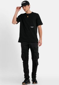 Zwarte katoenen t-shirt met logo print, gecombineerd met zwarte cargobroek met meerdere zakken. Draagt een zwarte pet en sneakers.