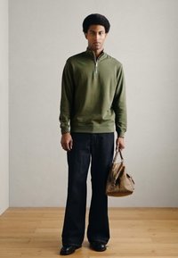 Roheline lukuga kaelusega kootud kampsun, tumedad lõhkuvad teksad, mustad kingad; hoiab pruuni kotti. Sile tekstuur ja vabaaja disain. Lihtne, neutraalne taust.