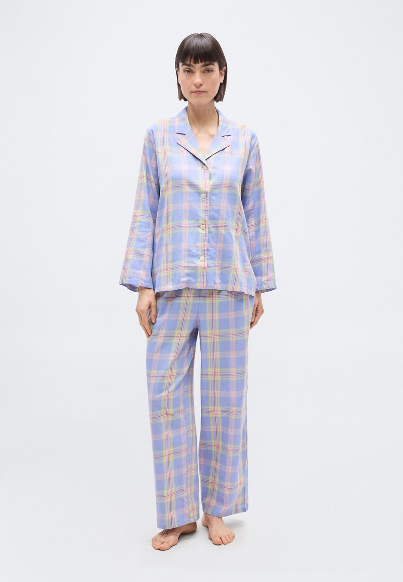 Γυναίκα όρθια, ξυπόλυτη, φορώντας πουκάμισο πιτζάμα με κουμπιά σε παστέλ μπλε, ροζ και κίτρινο καρό και φαρδιά παντελόνια που ταιριάζουν, με φόντο λευκό.