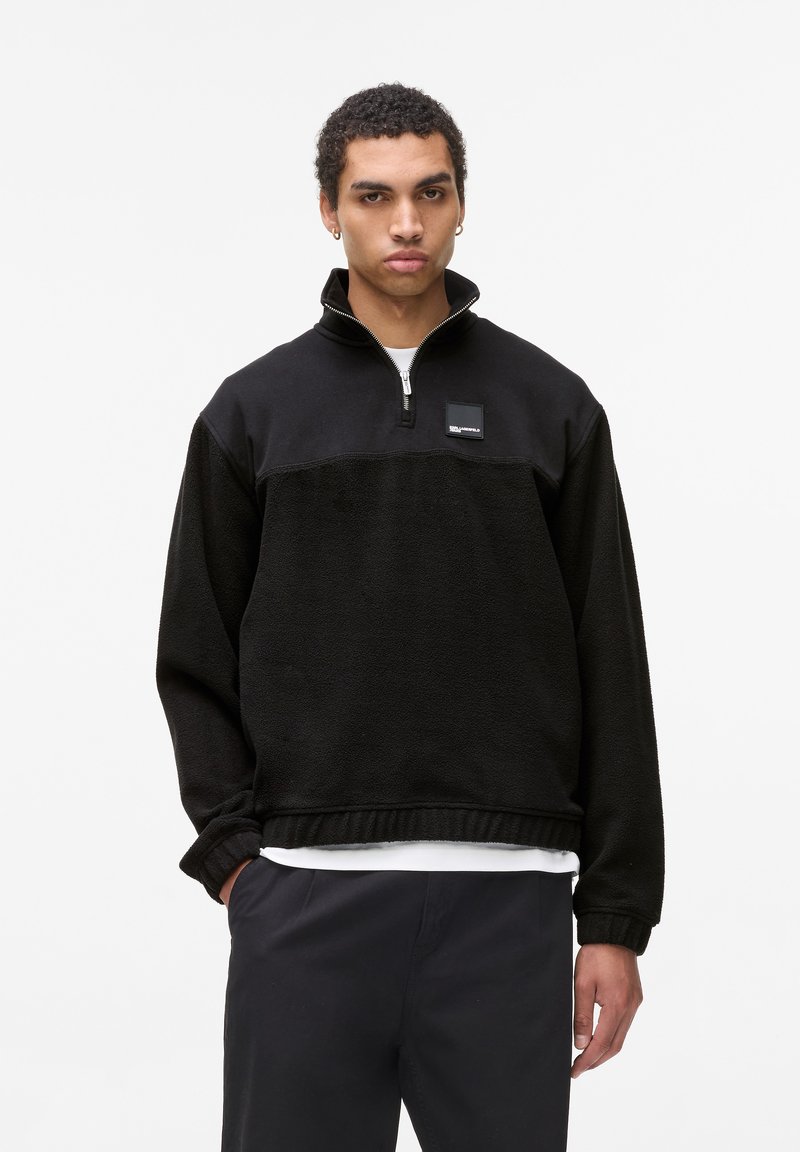 Giacca pullover in pile nero con collo a mezza zip, caratterizzata da un pannello superiore liscio a contrasto e un'etichetta del marchio tessuta sul petto.