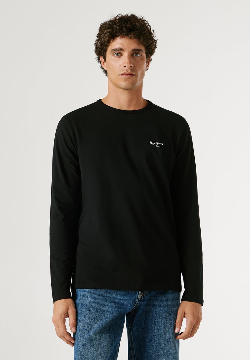 Mladi moški s kodrastimi lasmi, oblečen v črno majico z dolgimi rokavi in modre kavbojke, stoji pred enobarvnim svetlim ozadjem.