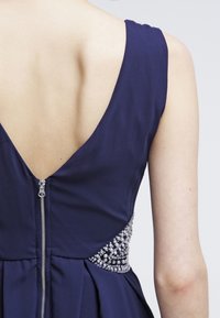 Rückansicht eines navyblauen ärmellosen Kleides mit einem tiefen V-Ausschnitt, silbernem Reißverschluss und verziertem Seitendetail an einer hellhäutigen Person.