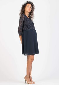 Navyblaues Kleid mit einem Spitzenoberteil, ¾ Ärmeln und einem fließenden Chiffonskirt. Das Design ist tailliert und eignet sich für Umstandsmode.