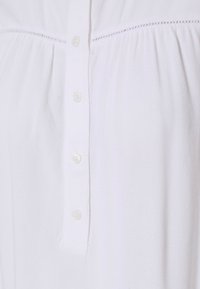 Camicia bianca con bottoni, caratterizzata da un tessuto strutturato, tre bottoni e un design plissettato sotto la yoke. Stile semplice e minimalista.