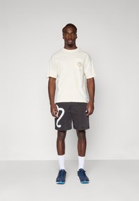 Lyse grå t-skjorte med kort erm og brystlomme, sammen med svarte shorts med et stort hvitt tall 2-design. Blå treningssko og hvite sokker.