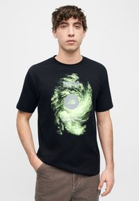 Νέος άνδρας με σγουρά μαλλιά που φοράει μαύρο μπλουζάκι με μια πράσινη γραφική σχεδίαση με στροφές και κείμενο που λέει "Running since 1906 New Balance Athletics Inc."