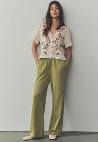 Cardigan crème en tricot avec broderies florales, associé à un pantalon large à rayures vertes. Les chaussures sont des ballerines couleur chair.