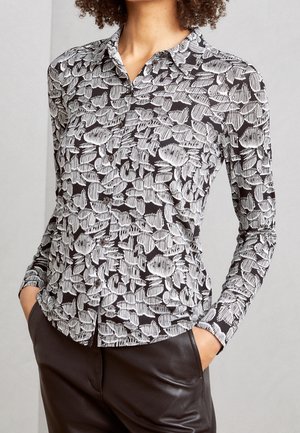 Femme portant une chemise cintrée à manches longues noire et blanche avec un motif floral abstrait et un pantalon en cuir foncé.