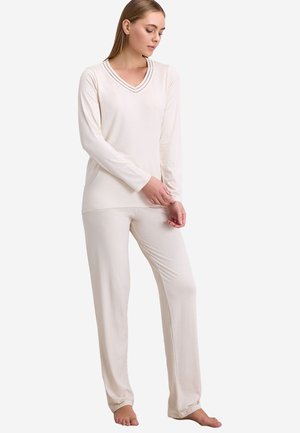 Vrouw staat op blote voeten, draagt een crèmekleurige top met lange mouwen en V-hals en bijpassende losse broek tegen een witte achtergrond.