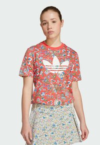 Röd blommig t-shirt med korta ärmar och vita logodetaljer, matchad med en vit kjol med färgglada blommönster och veck.