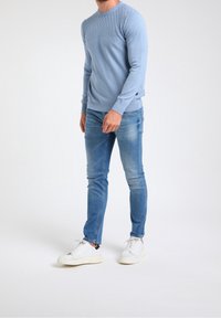 Mężczyzna ubrany w jasnoniebieski teksturowany sweter, wyblakłe niebieskie dżinsy i białe trampki, stojący na jednolitym białym tle.