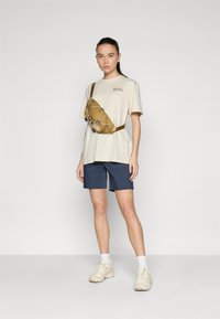 Béžové tričko s logom, námornícke šortky, hnedá ľadvinka, v kombinácii so svetlými teniskami a bielymi ponožkami. Jednoduchý, neformálny outfit na von.