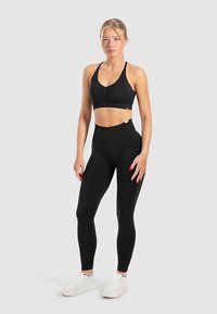Zwarte sportbh met geplooide voorkant, gecombineerd met zwarte leggings met hoge taille. Model draagt witte sportschoenen. Eenvoudig ontwerp, elastische stof.