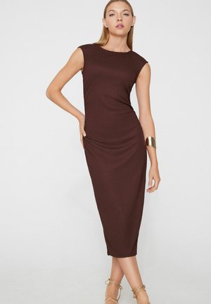 Femme portant une robe midi marron ajustée et sans manches, avec un bracelet manchette en or, posant avec une main sur la hanche et les jambes croisées sur fond blanc.