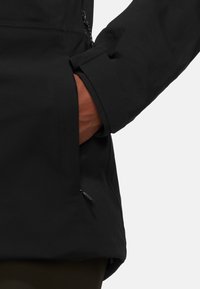 Svart ytterkläder med en slät textur, som har en sidoficka och justerbara ärmslut. Materialet verkar vara väderbeständigt.