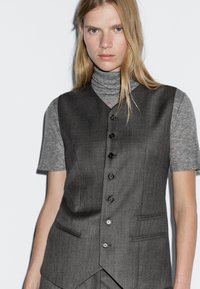 Gilet grigio fitted con scollatura a V, sei bottoni neri e due tasche laterali. Indossato sopra una maglietta a maniche corte grigia con collo alto.