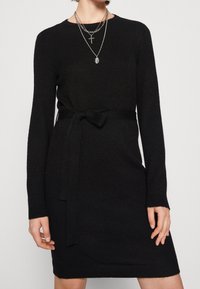 Abito nero in maglia con maniche lunghe, scollatura rotonda e una cintura di tessuto legata in vita. Texture liscia, design minimal.