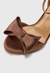 Sandale en satin marron avec un grand nœud décoratif à l'avant, design ouvert à l'avant et bride de cheville. Texture lisse et léger talon.