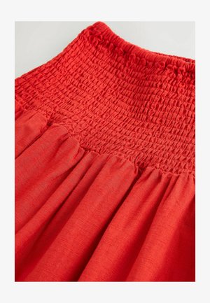 Punainen smokkihame, jossa on teksturoitu elastinen vyötärö ja pleatattu kangas, valmistettu kevyestä materiaalista liikkumisen helpottamiseksi.