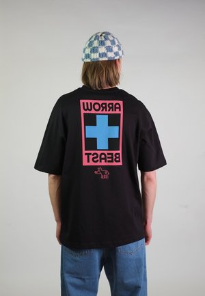 Persona che indossa una maglietta nera con croce blu bordata di rosa e testo rovesciato "ARROW BEAST" sulla schiena, cappello di maglia a quadri blu e jeans blu.