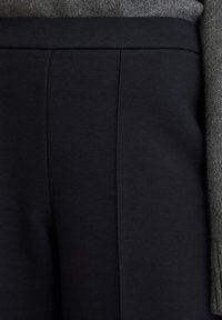 Zbliżenie na ciemnogranatowe spodnie z płaskim paskiem, noszone z szarym swetrem, którego rękaw widoczny jest z boku.