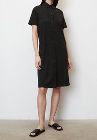 Svart kortärmad skjortklänning med krage, slättyg, knä-length nederkant och sidslits, tillsammans med platta svarta sandaler.