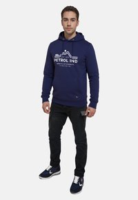 Felpa con cappuccio blu navy con lacci, testo bianco di marca e grafica di montagne. Abbinata a pantaloni neri e sneakers blu navy con lacci bianchi.