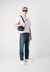 Sac bandoulière noir avec une étiquette bleue, porté par une personne en t-shirt blanc et jeans bleus, se tenant devant un fond clair.